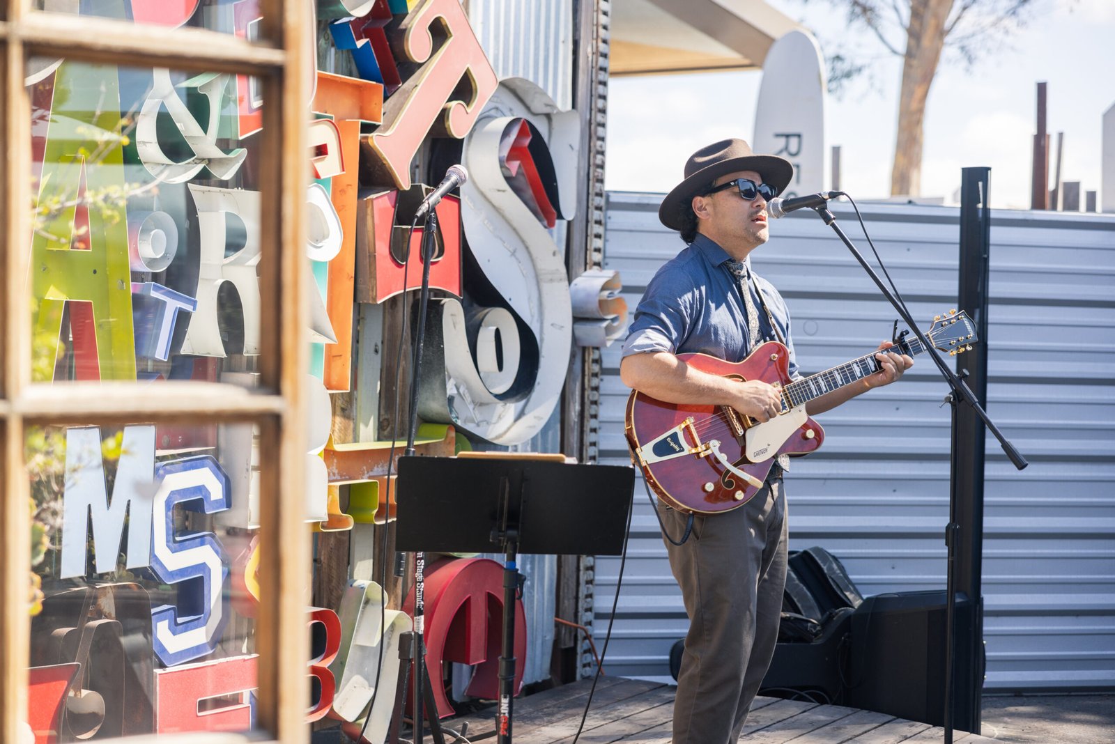 A man wearing a hat and sunglasses performs live music on a guitar outdoors, standing near a wall with large, colorful metal letters behind him; a microphone and music stand are in front of him.