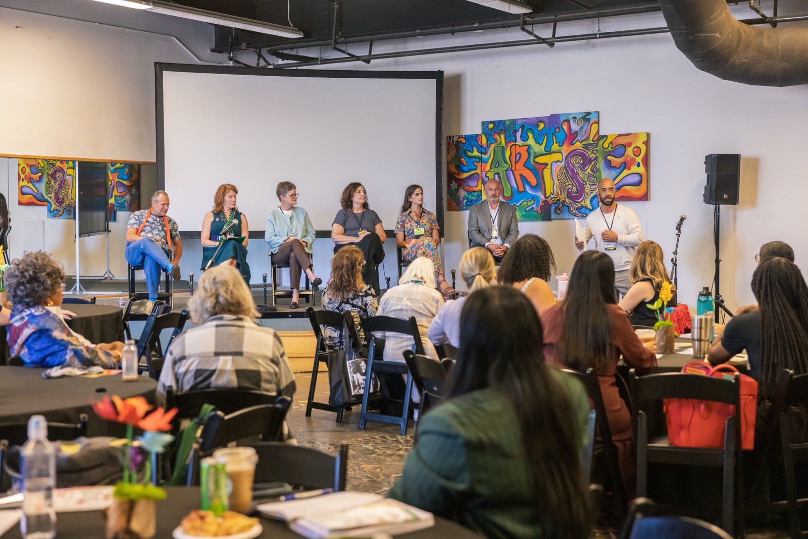 A panel discussion with six panelists sitting on chairs on a stage in front of a white screen, with a moderator standing at a podium and addressing an audience seated at round tables. The stage is decorated with colorful artwork. The audience is comprised of individuals, some taking notes and drinking beverages.