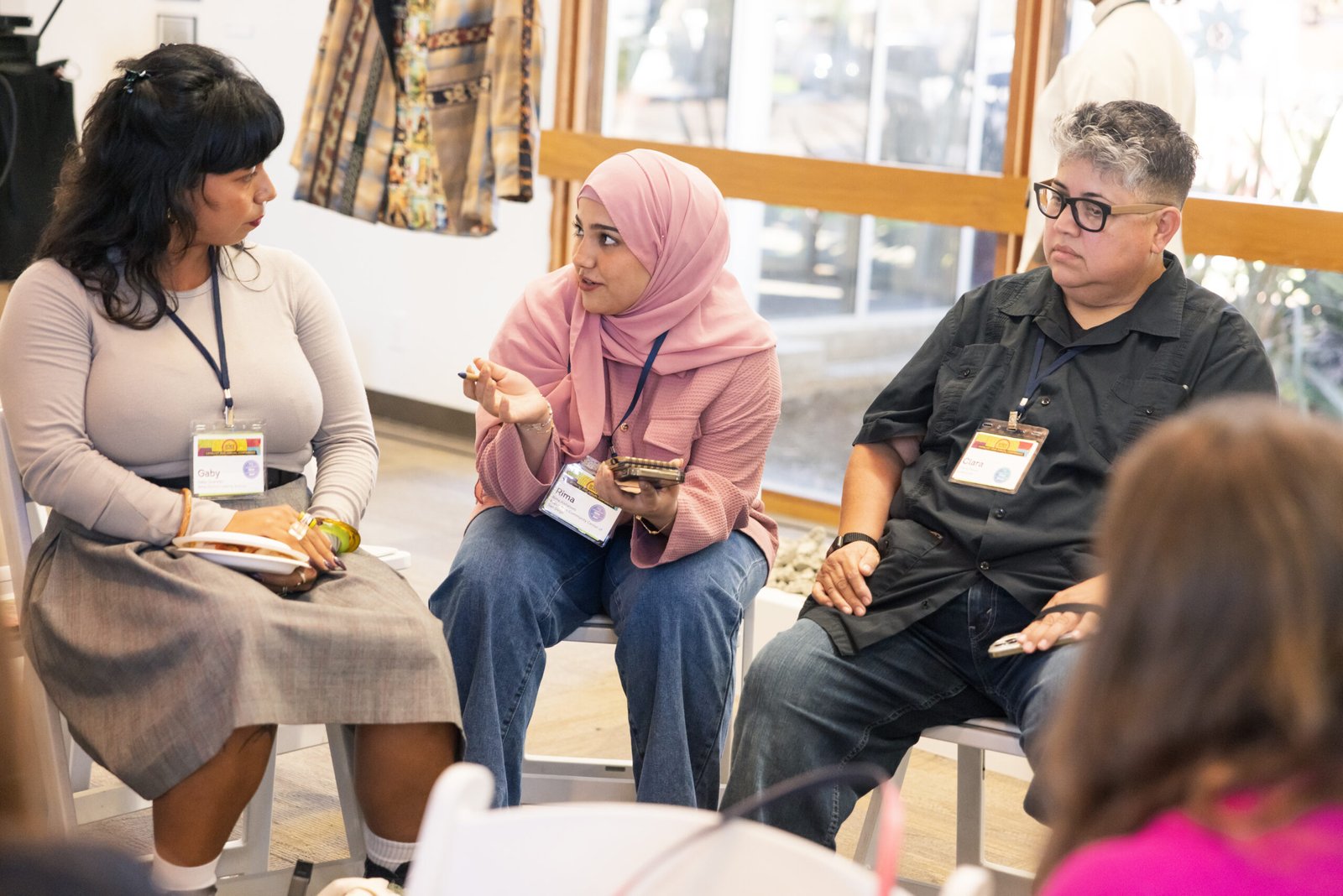 Three people, identified by name badges, sit in chairs in a light-filled room, possibly at a conference or workshop. The person in the middle, wearing a pink hijab and holding a pen and notebook, appears to be speaking to the group. The other two people, one on each side, are listening. A partially visible figure is in the foreground.