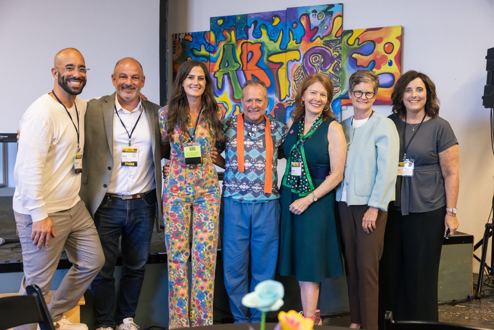 Seven people posing for a group photo on a stage, some wearing speaker badges, with a colorful abstract artwork in the background.
