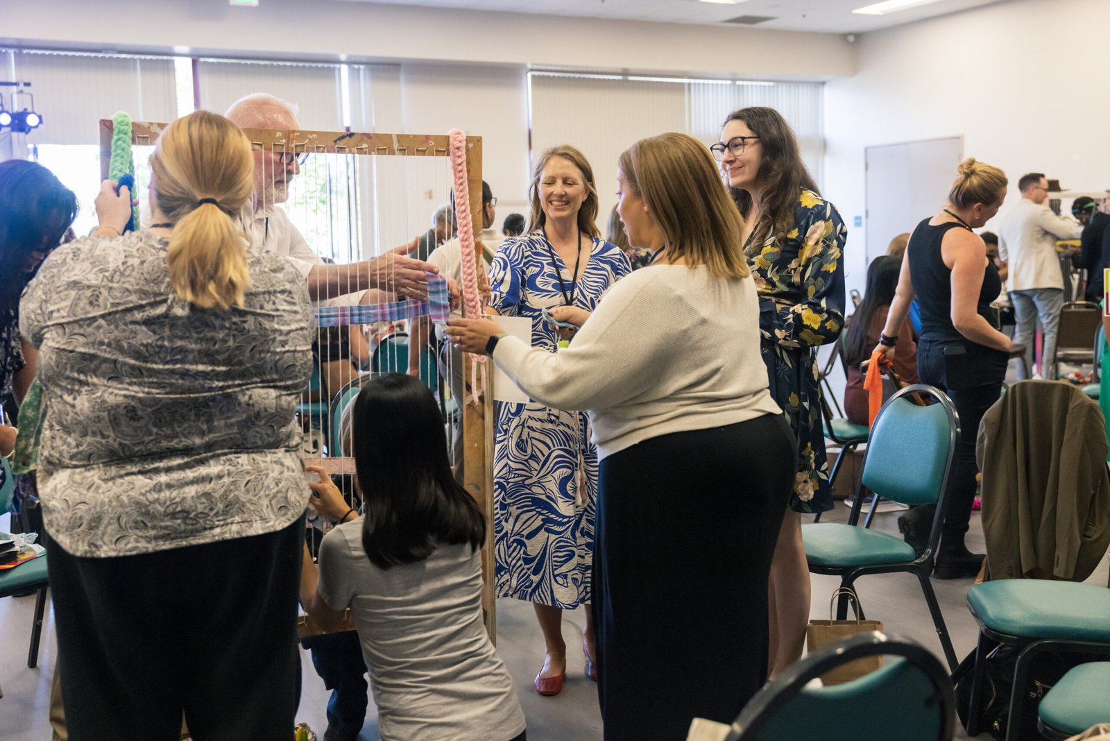 A group of people working on a weaving project, utilizing a large wooden frame with vertical threads. Various individuals are engaged in weaving and observing, with a woman in a floral dress and a man with glasses are interacting near the frame. The setting appears to be a conference or workshop room.