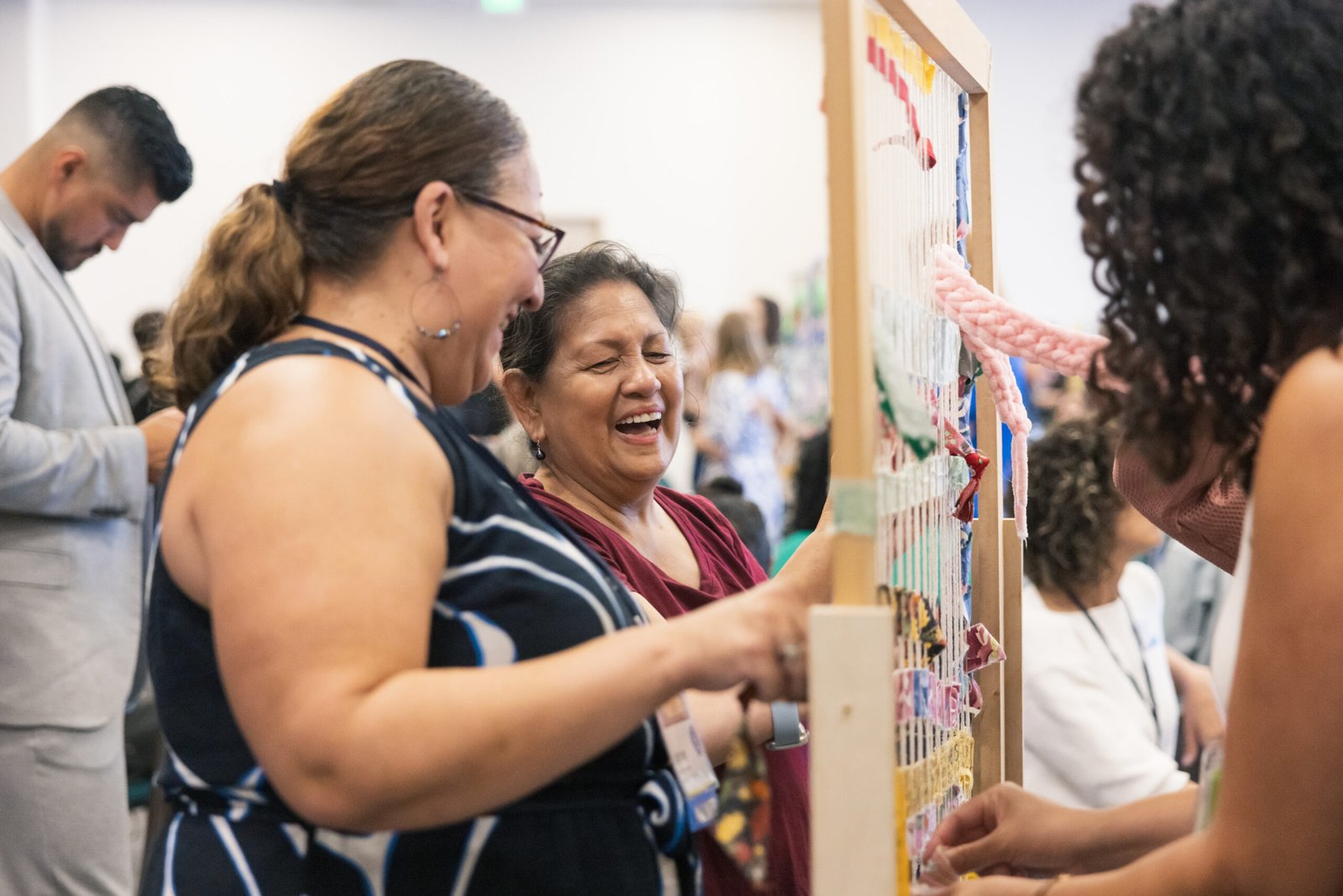 Two women laughing while working on a weaving loom, with various colorful fabrics and yarns attached. Other people are in the background.
