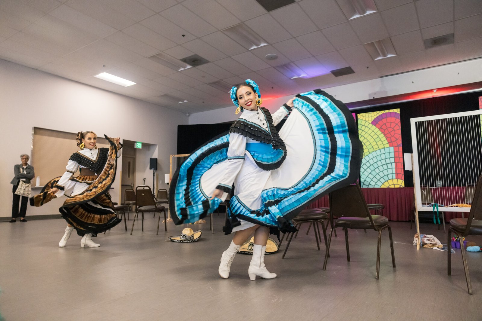 Two women in traditional Mexican dresses dancing in an open space, each with a wide skirt billowing outwards. The woman in focus wears a white dress with a blue and black design, adorned with matching accessories in her hair and white boots, while the other dancer wears a similar style dress in brown and black. A sombrero sits on the floor. An older woman watches in the background and a stage is partially visible.