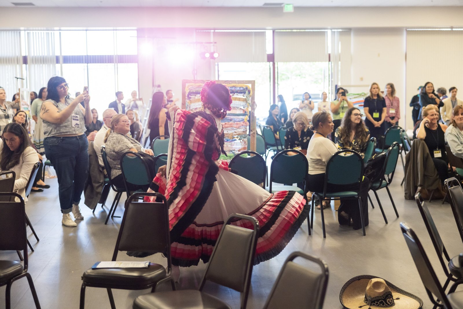 A woman in a vibrant, tiered, red and white traditional Mexican dress and headdress dances in a large auditorium in front of an audience, many of whom are seated and taking photos.