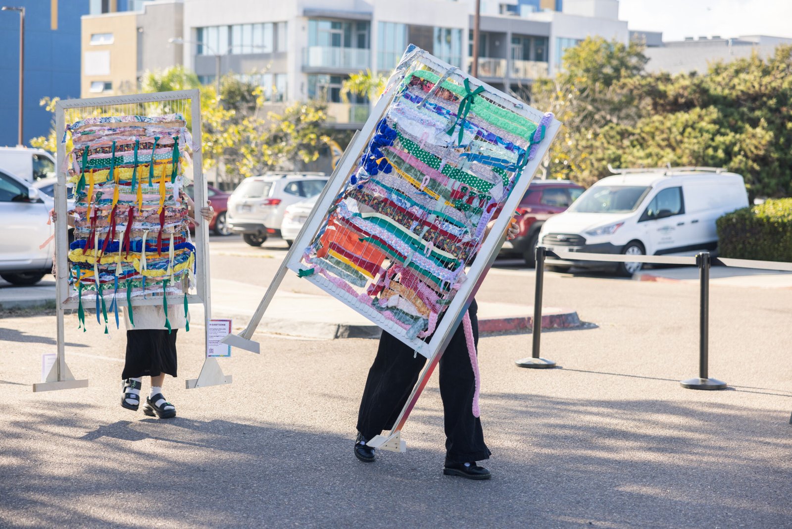 Two people walking on asphalt, each carrying a large, rectangular frame filled with colorful fabric strips, resembling woven artwork, with a building and parked vehicles in the background.