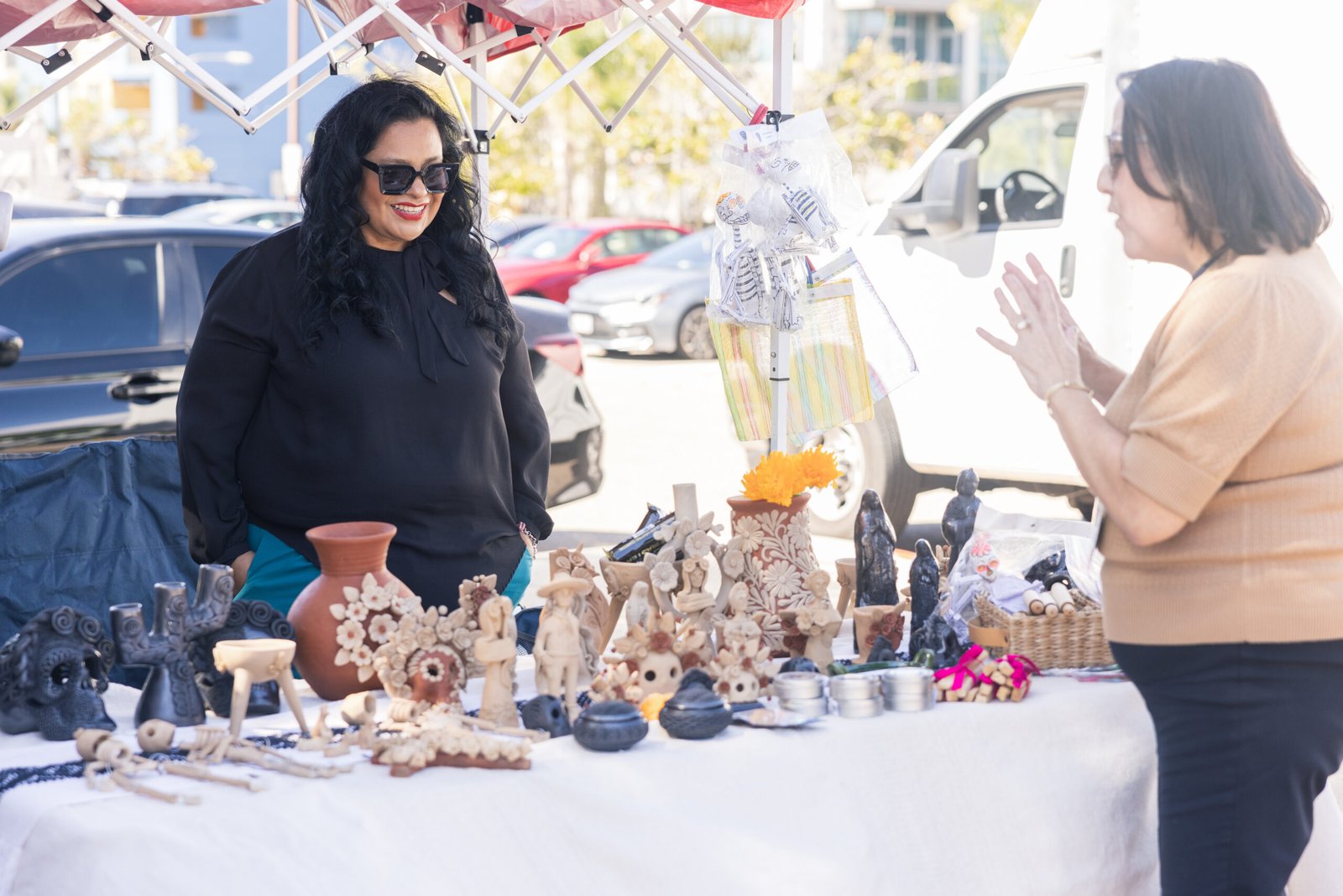 Two women at a craft fair stand: One, with sunglasses and black attire, stands behind a table laden with Day of the Dead themed pottery and ornaments; the other, wearing a beige top and black skirt, gestures with open hands towards the display. A white van is parked nearby, and cars are in the background under a red and white canopy.
