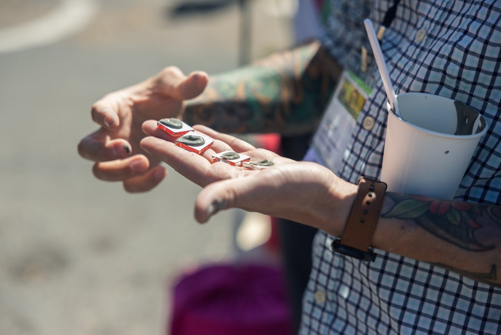 Person holding multiple, small, rectangular glass tiles topped with grey substance in their hand. The person has several tattoos on their arms and wears a brown watch on their wrist. A paper cup with a straw sits on the person's arm. The person wears a plaid shirt.
