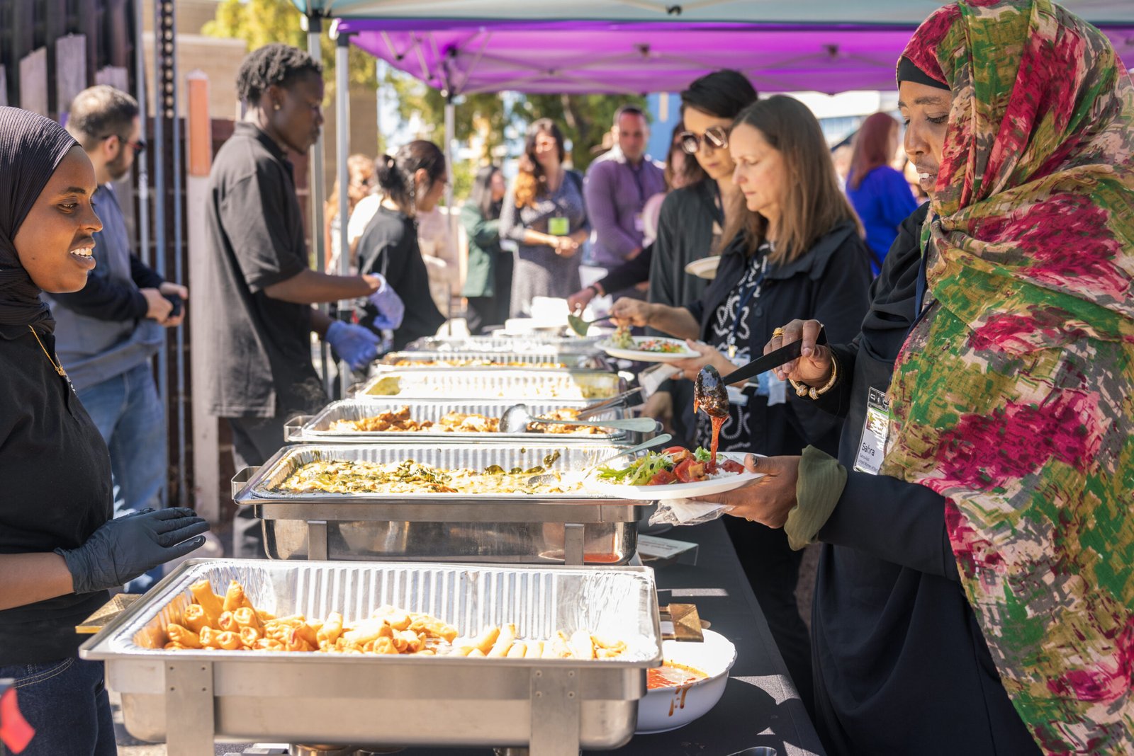 A diverse group of people serve themselves food from buffet trays at an outdoor event. One woman in a colorful patterned hijab and black clothing serves food onto a white plate, as others queue behind her to serve themselves. In the foreground, other people are seen serving food and wearing protective gloves. A variety of cooked dishes are presented in silver chafing dishes under a purple tent.