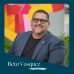 Beto Vasquez, a smiling man with glasses and a goatee, wearing a navy blazer and black t-shirt, stands in front of a colorful mural.