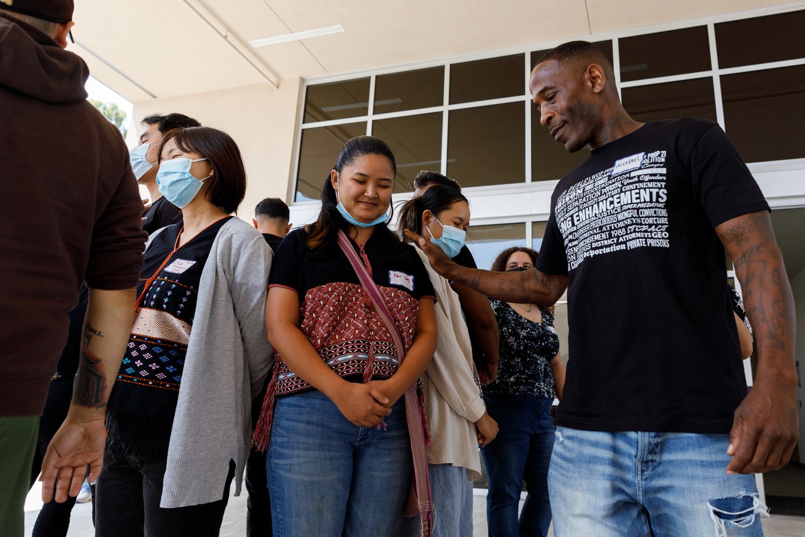 A group of people, most wearing face masks, standing in line and facing forward outside a building. A man in a black t-shirt with "ENHANCEMENTS" in large white text and smaller text with legal terms touching the shoulder of a woman wearing a mask and a patterned top. A name tag reads "Paw Say." Another name tag reads "Nao Kalsberg 1993." A name tag reads "Michael Camper."
