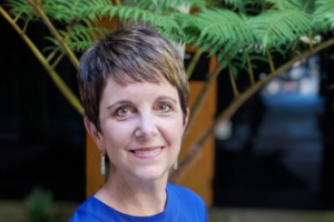 A photo of Nancy Jamison from the shoulders up. She is wearing a bright blue top and has a green plant behind her.