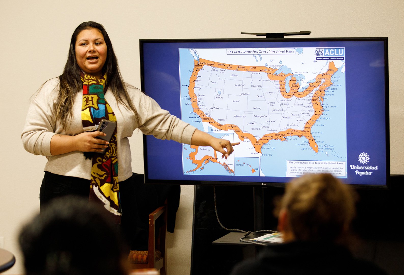 A woman with dark hair, wearing a patterned scarf with a "H" graphic, stands in front of a screen displaying a map of the United States with highlighted areas and the title "The Constitution-Free Zone of the United States," alongside the ACLU logo. The text on the screen includes city names, geographic markings, and the statement: "Nearly 2 out of 3 Americans (197.4 million people) live within 100 miles of the US land and coastal borders, according to 2007 figures from the Census Bureau." The screen also features the "Universidad Popular" logo. The woman is gesturing with her hand towards the map. Several blurred individuals are in the foreground, indicating an audience or group setting.