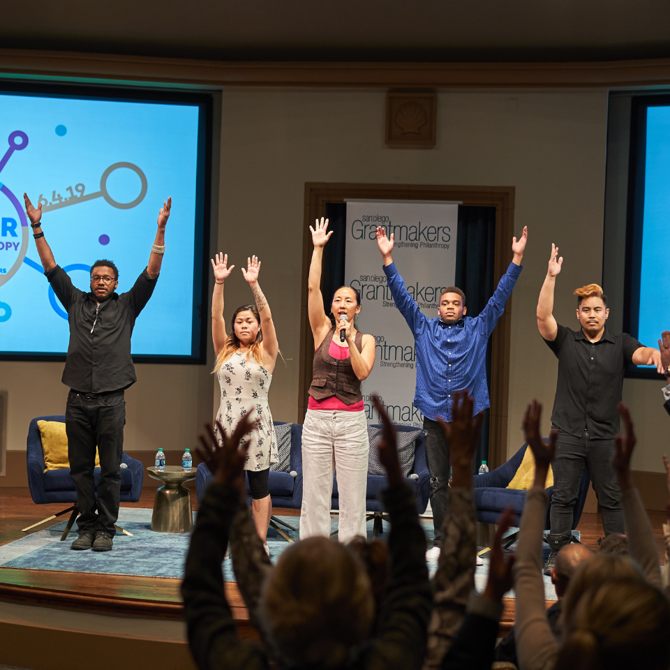 Five diverse individuals stand on a stage with arms raised in unison, likely engaging with an audience whose raised hands are visible in the foreground. A woman holds a microphone. The backdrop includes two screens displaying graphics, and a banner reading "San Diego Grantmakers."