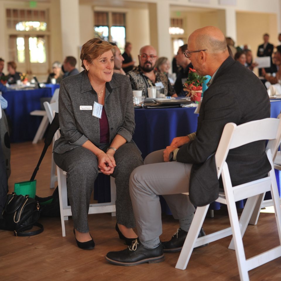 Two individuals, a woman and a man, seated and engaged in conversation at a conference or event, with others visible in the background seated at tables with blue tablecloths. The woman, wearing a gray suit and a name tag, is attentively looking at the man, who is wearing a dark jacket, gray pants, and glasses. Both are seated in folding chairs.