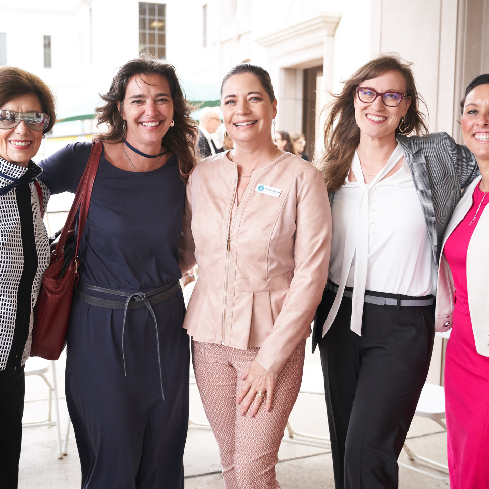 Five smiling women posing together outdoors, with the woman in the center wearing a light pink jacket and patterned pants.