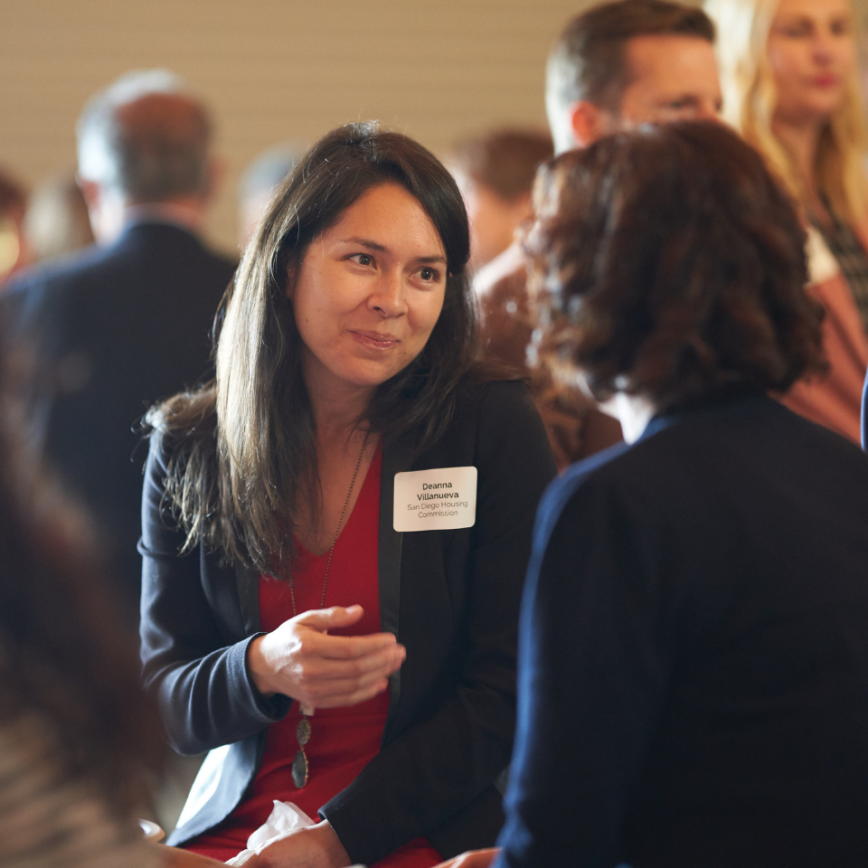 Deanna Villanueva, wearing a name tag identifying her as a member of the San Gabriel Valley Housing Commission, speaks animatedly to another person at an event, gesturing with her right hand. She wears a black blazer over a red dress and a pendant necklace.