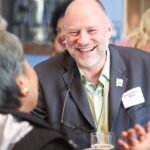 A smiling, middle-aged bald man in a suit jacket and glasses hanging from his shirt, talking and gesturing at an event; a name tag reads "Tomas Herrera-Mishler."