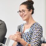 Image Preview A smiling woman with glasses and a bun, wearing a patterned blue and white blouse, holding a microphone and gesturing during a panel discussion.