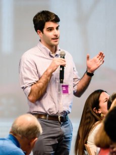 A young man with dark hair, wearing a light blue checked shirt and belt, holding a microphone, gesturing with his right hand as he speaks to an audience; a purple name tag is on his chest.