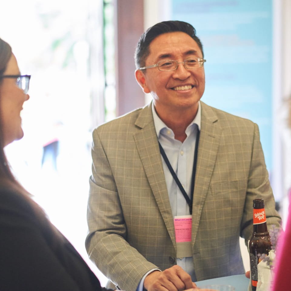 Smiling Asian man in a plaid blazer and glasses, wearing a lanyard and nametag, talking to another person out of frame, with a bottle of Shock Top beer on a table nearby.